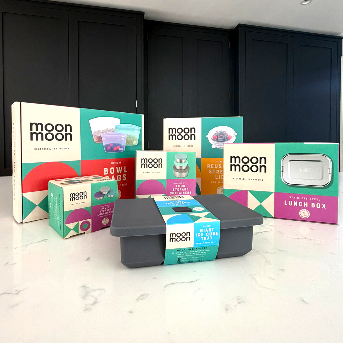 A collection of Moon Moon branded food storage products arranged on a white marble countertop. Visible items include a gray silicone ice cube tray, multiple colorful stainless steel and silicone storage containers, and boxed sets such as free-standing silicone bowl bags, silicone stretch lids, stainless steel lunchbox and sauce pots.