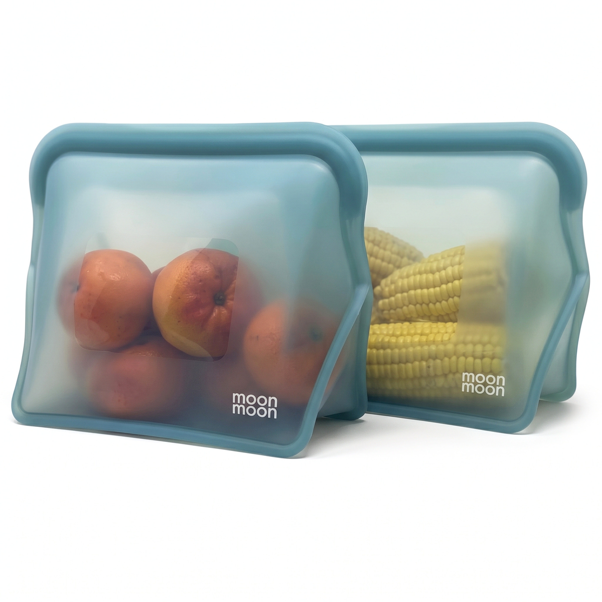 Two moonmoon reusable silicone food storage bags with wide stand-up bases and rounded corners in a muted blue-green color, shown on a white background. One bag contains red apples, and the other contains yellow corn.